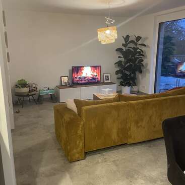 Uma sala de estar aconchegante com um sofá amarelo mostarda, uma TV exibindo uma lareira virtual, plantas domésticas, decoração moderna e grandes janelas com vista para uma área externa.