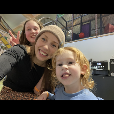 Três pessoas sorridentes, incluindo duas crianças e uma mulher com um gorro, posam para uma selfie alegre em um playground com uma máquina de café atrás delas.