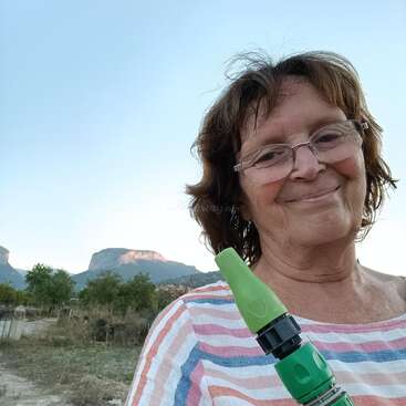 A imagem mostra uma mulher idosa com cabelo castanho curto, vestindo uma camisa listrada, em pé em frente a uma paisagem montanhosa, segurando um bico de mangueira verde.