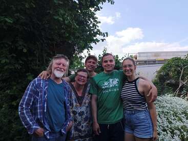 L'image représente un groupe de cinq personnes posant pour une photo dans un jardin, avec un ciel bleu serein et des nuages blancs en toile de fond.