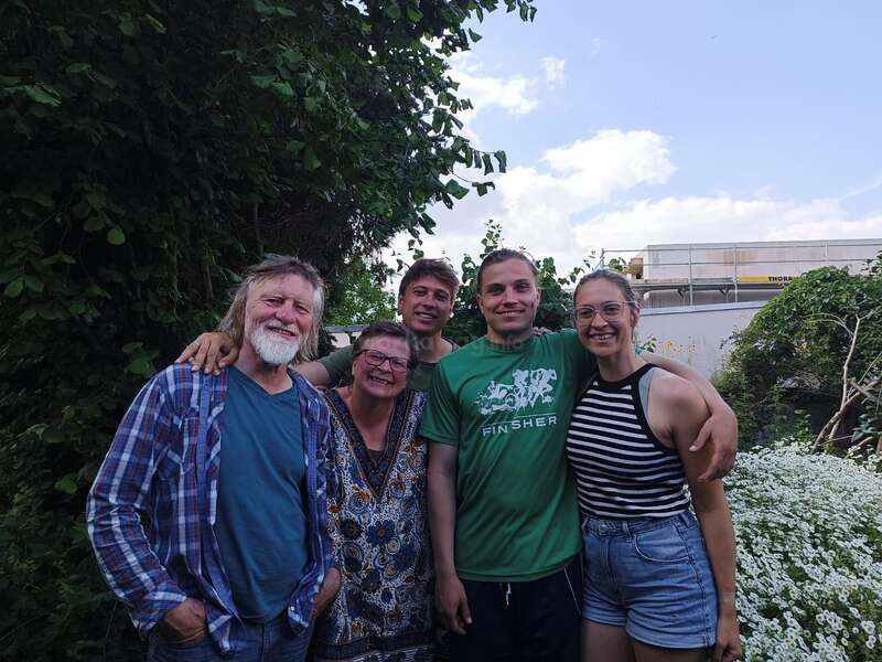 A imagem mostra um grupo de cinco pessoas posando para uma foto em um jardim, com um céu azul sereno e nuvens brancas como pano de fundo.