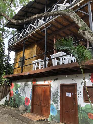 Auf dem Bild sieht man ein zweistöckiges rustikales Gebäude mit Holztüren, bunten Blumenwandbildern, weißen Geländern, Plastikstühlen und Tischen auf dem Balkon und viel Grün drumherum.