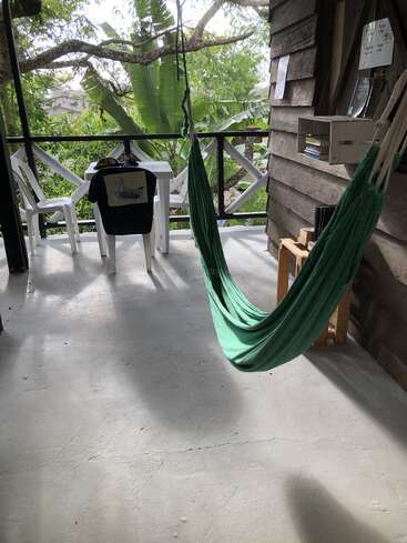 Eine gemütliche Veranda mit einer grünen Hängematte, weißen Plastikstühlen, einem kleinen Tisch, auf dem ein Hemd liegt, umgeben von üppiger tropischer Vegetation und Büchern.