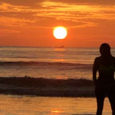 A silhueta de uma pessoa está na praia, olhando para um pôr do sol laranja intenso sobre ondas calmas, com um barco distante no horizonte.