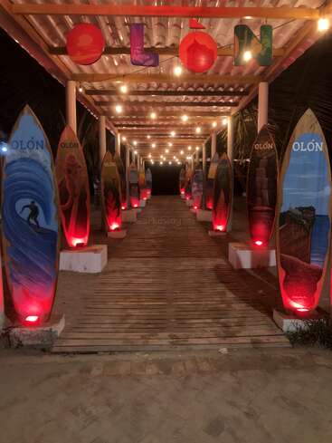 Eine Holzpromenade, die mit bemalten Surfbrettern geschmückt ist und nachts von roten Lichtern und Lichterketten beleuchtet wird, unter einem Schild mit der Aufschrift „OLÓN“. Strandatmosphäre, einladend.