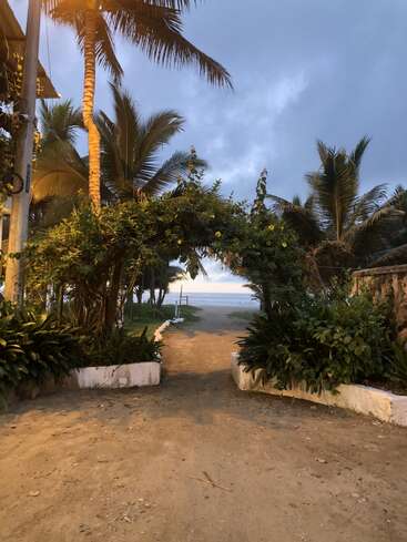 Ein sandiger Weg, umgeben von viel Grün und Palmen, führt durch einen Torbogen zu einem ruhigen Strand und dem stillen Meer unter einem wolkenverhangenen blauen Himmel in der Abenddämmerung.