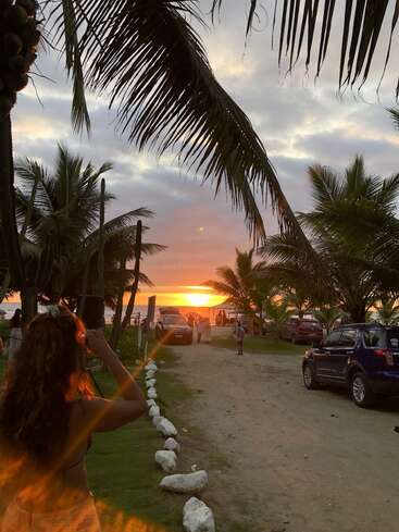 Uma mulher contempla um pôr do sol deslumbrante sobre o oceano, emoldurado por palmeiras, carros e pessoas, criando uma atmosfera tropical e serena de praia.
