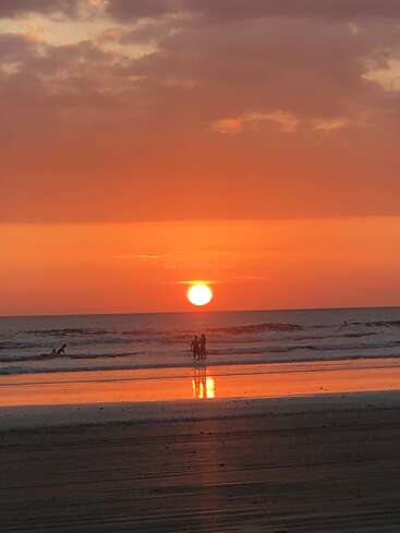 Ein leuchtend orangefarbener Sonnenuntergang über dem Meer, mit Menschen, die als Silhouetten in den flachen Wellen stehen. Die Spiegelungen schimmern auf dem nassen Sand und schaffen eine friedliche, ruhige Strandszene.