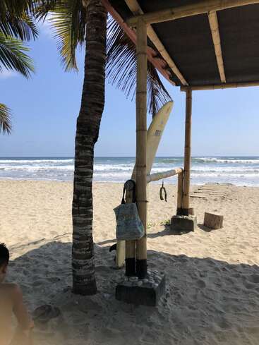 Eine ruhige Strandszene mit sandigem Ufer, Bambushütte, Surfbrett, Palme, einer hängenden Tasche, strahlend blauem Himmel, sanften Wellen und einer Person, die in der Nähe sitzt.