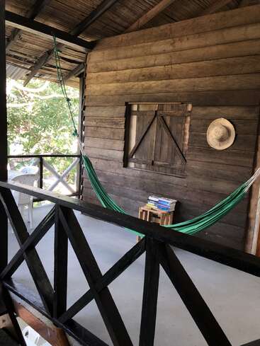 Eine gemütliche Holzveranda mit einer grünen Hängematte, rustikalen Wänden, einem Strohhut, einem kleinen Tisch voller Bücher und einem Plastikstuhl, die zum Entspannen einlädt.