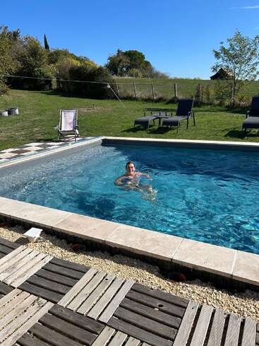 Una persona está nadando en una piscina al aire libre en un día soleado. Alrededor de la piscina hay sillas de jardín, césped verde, árboles y una terraza de madera.