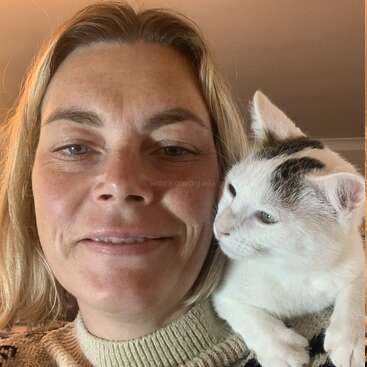 Una mujer de pelo rubio sonríe amablemente mientras un gatito blanco con marcas negras se sienta en su hombro, mirándola cariñosamente. Ambiente acogedor y cálido.