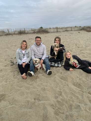 Una familia de cuatro miembros está sentada en una playa de arena con dos perros pequeños, sonriendo. Detrás de ellos hay una valla de madera y el cielo nublado, creando un ambiente relajado.