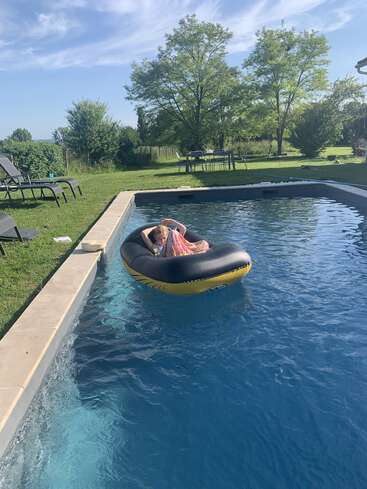 Un niño se relaja en una balsa hinchable amarilla en la piscina del patio trasero, rodeado de césped verde, tumbonas y árboles bajo un cielo azul.
