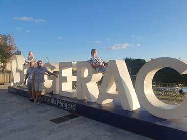 Cuatro personas posan alrededor de las grandes letras blancas "BERGERAC" en un soleado entorno al aire libre. Dos de pie en el suelo, dos sentadas en lo alto del letrero, sonriendo bajo el cielo azul.