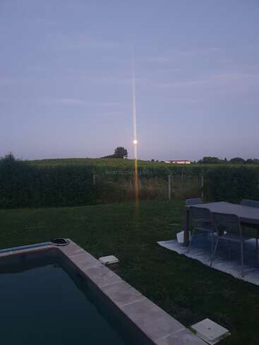 Una tranquila escena nocturna con una piscina, un comedor al aire libre sobre una alfombra, un patio cubierto de hierba, campos lejanos, setos y una brillante luna llena en el horizonte.