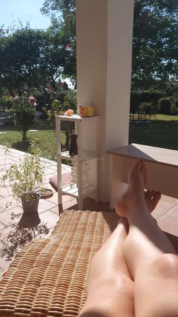 Una persona se relaja en un patio soleado, con las piernas estiradas en una silla de mimbre. Plantas en macetas, flores y vegetación de exterior crean un ambiente de jardín tranquilo y veraniego.