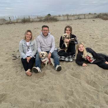 A family of four sits on a sandy beach with two small dogs, smiling. Behind them is a wooden fence and cloudy sky, creating a relaxed atmosphere.