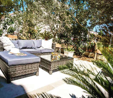 La imagen representa un sereno patio exterior con un sofá de mimbre, una mesa de centro y una exuberante vegetación, que evoca una sensación de relajación y tranquilidad.