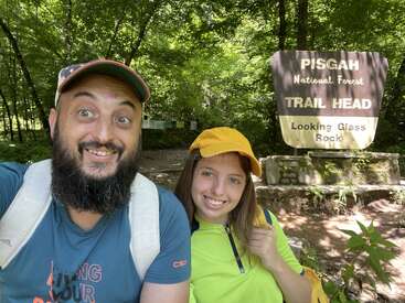 A man and girl smile at the Pisgah National Forest Trail Head for Looking Glass Rock, wearing hiking gear and backpacks, surrounded by lush green trees.
