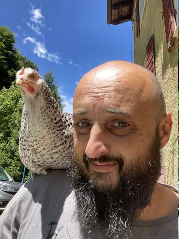 A bald man with a beard smiles while a speckled chicken perches on his shoulder. The background shows a sunny day, blue sky, and greenery.