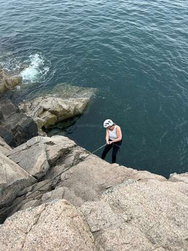 Jemand mit Helm seilt sich an einer felsigen Klippe am Meer ab und hält sich an einem Seil über dem tiefblauen Wasser fest, während in der Nähe Wellen brechen.