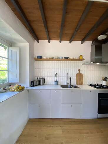 A bright, minimalist kitchen with white cabinets, wooden ceiling, and open shelf. Fruit, kitchen appliances, and utensils are neatly arranged, with sunlight streaming through a window.