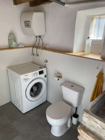 This image shows a small laundry bathroom with a washing machine, toilet, yellow towel, soap bottle, water heater, window, and a rustic stone sink.