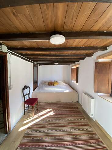 This cozy bedroom features a wooden beamed ceiling, white walls, a large bed with pillows, a striped rug, two windows, and a vintage chair.