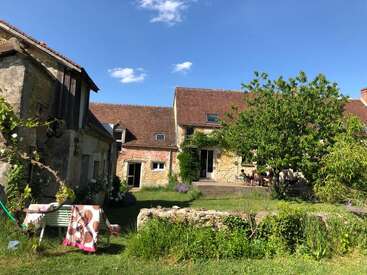 Das Bild zeigt ein charmantes, rustikales Steinhaus mit rotem Ziegeldach, umgeben von viel Grün, einem Hof, Gartenpflanzen und einem Tisch mit Stühlen im Freien.