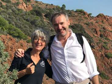 Un homme et une femme souriants posent ensemble en plein air, entourés de collines rocheuses rouges et de verdure. Tous deux ont l'air heureux, vêtus de tenues décontractées, profitant d'une journée ensoleillée pour faire une randonnée.