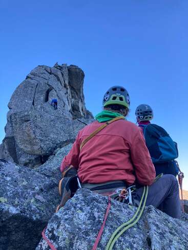Deux grimpeurs équipés de casques et de matériel d'escalade sont assis sur un terrain rocheux et se préparent à monter. Un autre grimpeur se trouve plus haut, sur une formation rocheuse abrupte et déchiquetée.