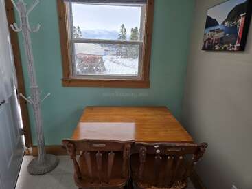 Un coin confortable avec une table en bois et deux chaises se trouve près d'une fenêtre. Dehors, la neige recouvre le paysage. Un porte-manteau et un tableau décorent la pièce.