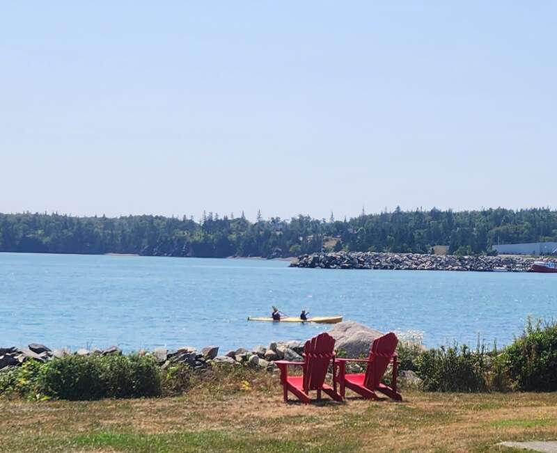 Une scène paisible au bord d'un lac avec deux chaises Adirondack rouges sur l'herbe, un rivage rocheux, deux kayakistes dans l'eau, avec des arbres et un ciel bleu en arrière-plan.