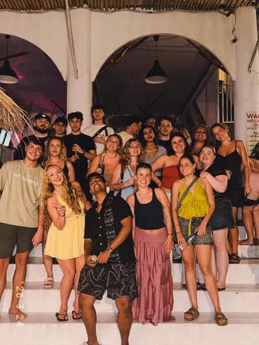 A group of young adults pose together on white stairs, smiling and enjoying a night out. Some hold drinks, dressed in casual summer clothing, looking happy.