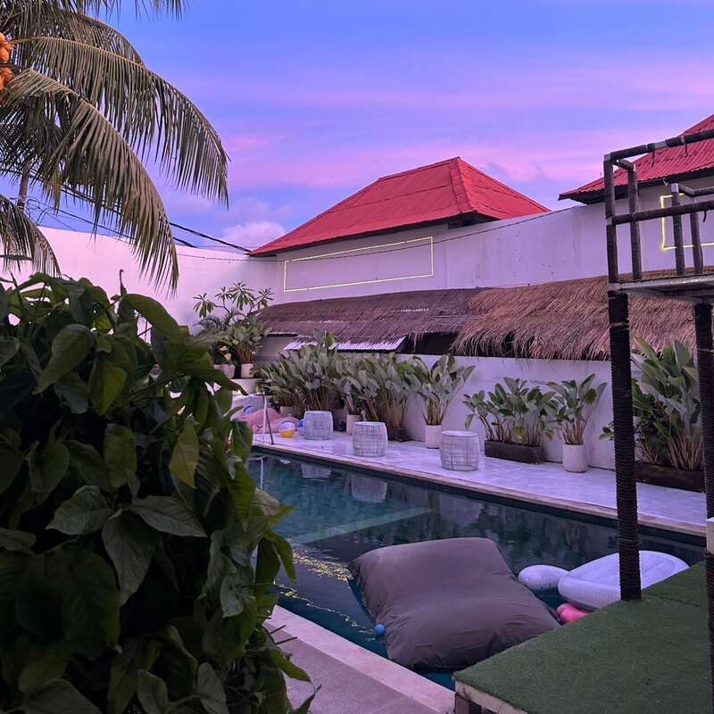 A serene poolside scene at sunset with lush greenery, floating pool toys, red-roofed buildings, palm trees, and a purple-pink sky creating a tropical ambiance.