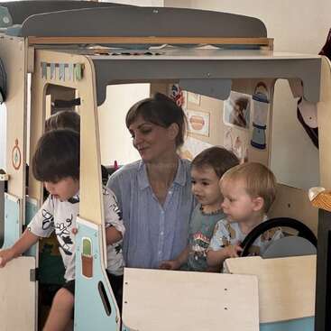 A woman and three young children are playing together inside a wooden play vehicle, smiling and engaged in imaginative play. The scene feels joyful and nurturing.