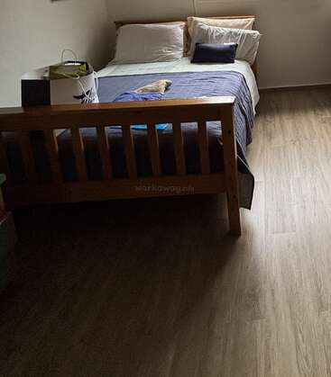 A cozy bedroom with a wooden bed frame, blue and white bedding, three pillows, a small pillow, a shopping bag, and wooden flooring, warmly lit.