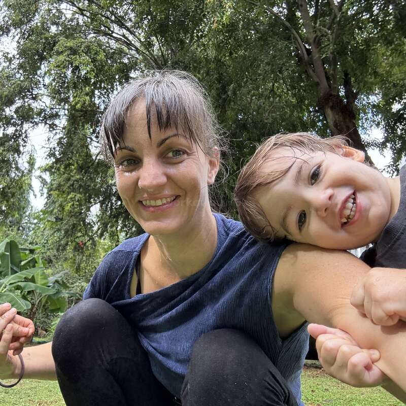 A woman and a child smile joyfully outdoors, sitting on grass with lush green trees in the background. They look happy, playful, and enjoy each other’s company.