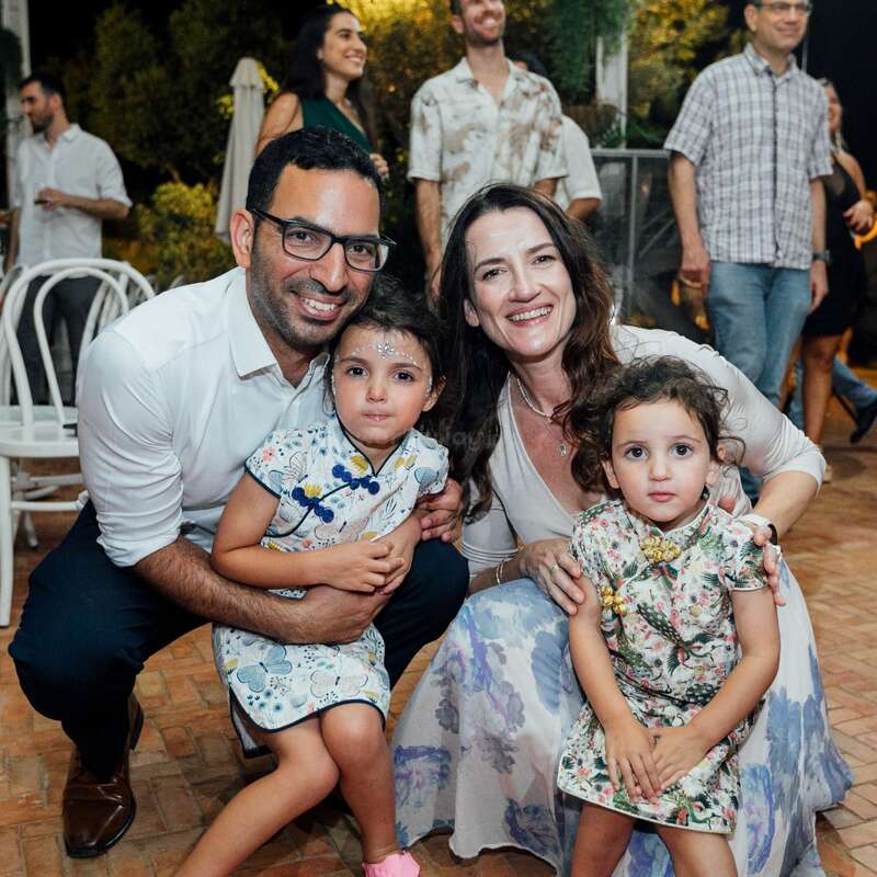 A smiling family of four poses together at a lively event. The parents crouch beside their two young daughters, surrounded by other people in a festive atmosphere.