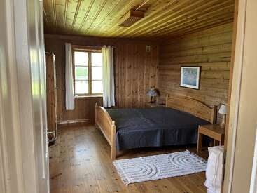 Esta imagen muestra un acogedor dormitorio de madera con una cama grande, una colcha negra, dos mesitas de noche, una ventana con cortinas blancas, cuadros en la pared y una alfombra blanca.