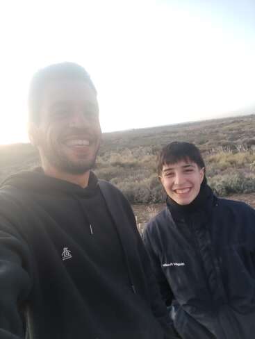 Duas pessoas estão sorrindo ao ar livre, vestidas calorosamente com jaquetas escuras. O plano de fundo mostra uma paisagem natural com arbustos sob um céu claro e brilhante. A atmosfera é alegre.