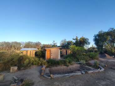 Dieses Bild zeigt kleine, rustikale Gebäude aus natürlichen Materialien, umgeben von Grünflächen. Der Himmel ist klar, und die Szene wirkt ruhig und ländlich.