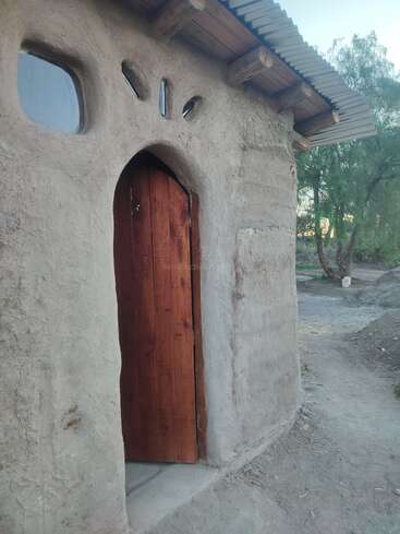 Dieses Bild zeigt ein rustikales, erdverbundenes Gebäude mit einer Holztür und organisch geformten Fenstern. Die Umgebung wirkt natürlich, mit Bäumen und einem Feldweg in der Nähe.