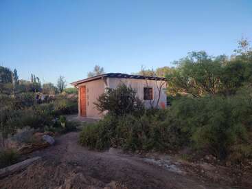 Dieses Bild zeigt ein kleines, bescheidenes Haus, das von grünen Sträuchern und Bäumen umgeben ist und zu dem ein unbefestigter Weg unter einem klaren blauen Himmel führt.
