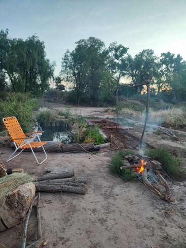Uma cena tranquila ao ar livre mostra uma cadeira de gramado laranja perto de um pequeno lago, cercada por troncos, uma fogueira modesta e uma vegetação exuberante sob um céu sereno.