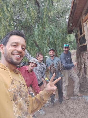 Cinco pessoas sorriem ao ar livre, posando para uma selfie em grupo perto de uma estrutura rústica de madeira. Elas parecem estar trabalhando juntas, com as mãos e as roupas cobertas de lama, celebrando o trabalho em equipe.
