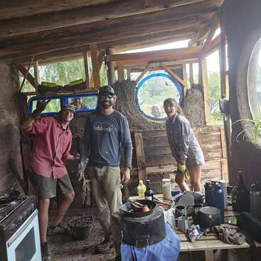 Trois personnes sourient à l'intérieur d'une structure rustique, partiellement construite, avec des poutres en bois et des murs en terre. Des ustensiles de cuisine encombrent une table et la lumière naturelle éclaire cette scène animée.