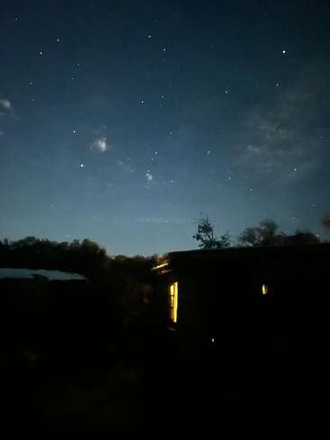 Um céu noturno escuro e tranquilo, repleto de estrelas cintilantes. Uma casa aconchegante abaixo tem uma luz quente brilhando através de sua janela, cercada por árvores e colinas em silhueta.