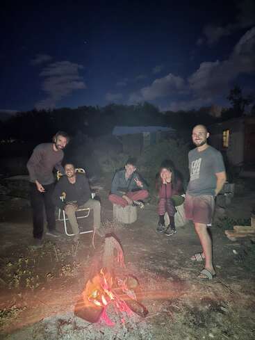 Cinco pessoas estão sentadas e em pé ao redor de uma fogueira à noite, sob um céu estrelado. Elas estão sorrindo, relaxadas e aproveitando o fogo quente e brilhante.